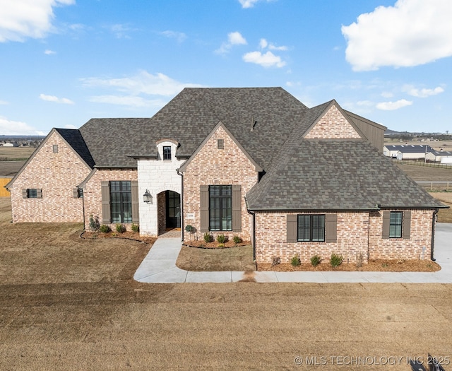 french country inspired facade with a shingled roof, a front lawn, and brick siding