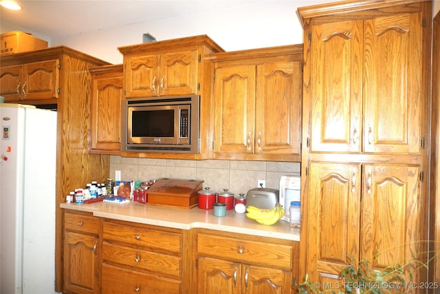 kitchen featuring tasteful backsplash, brown cabinets, light countertops, stainless steel microwave, and freestanding refrigerator