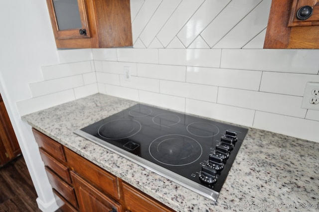 room details featuring light stone countertops, black electric stovetop, brown cabinetry, backsplash, and glass insert cabinets