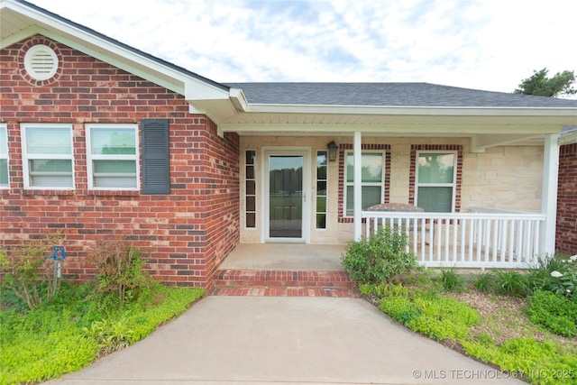 property entrance featuring brick siding, covered porch, and roof with shingles