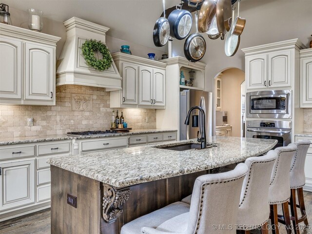 16321 E 48th Pl, Tulsa, OK 74134 - kitchen with a kitchen island with sink, a sink, custom range hood, arched walkways, and appliances with stainless steel finishes