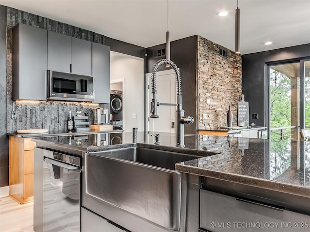 kitchen featuring stacked washer and dryer, dark stone counters, stainless steel microwave, backsplash, and a sink