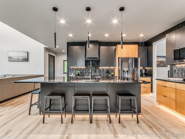 kitchen featuring wall chimney exhaust hood, a kitchen bar, modern cabinets, and stainless steel refrigerator with ice dispenser