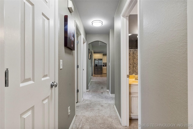 hall with a sink, a textured wall, baseboards, arched walkways, and light colored carpet