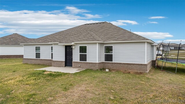 back of property with a trampoline, a yard, brick siding, a shingled roof, and a patio area