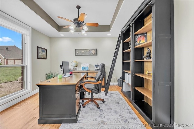 office featuring a tray ceiling, baseboards, a ceiling fan, and light wood finished floors