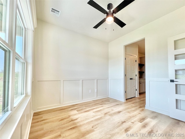 empty room with visible vents, a wainscoted wall, light wood-type flooring, a decorative wall, and a ceiling fan