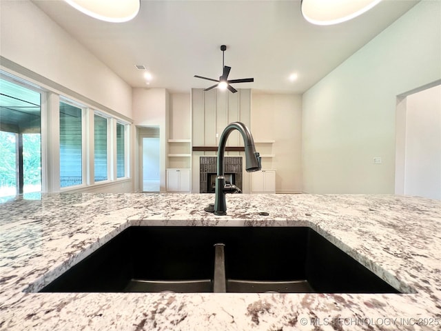 kitchen featuring a sink, a fireplace, a ceiling fan, visible vents, and light stone counters
