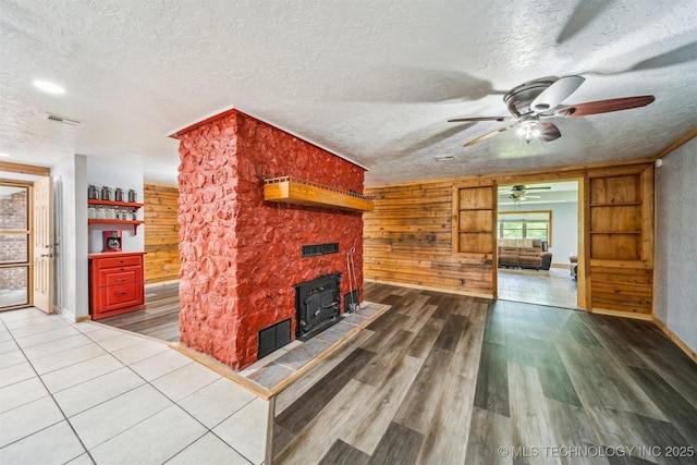 unfurnished living room with a textured ceiling, a wood stove, ceiling fan, visible vents, and wood finished floors