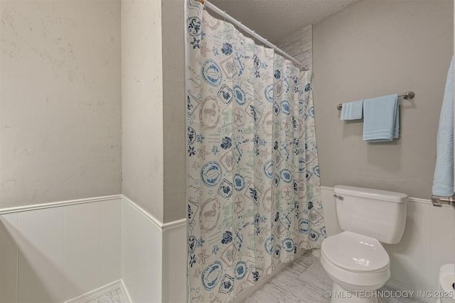 full bathroom featuring a textured ceiling, curtained shower, a wainscoted wall, toilet, and marble finish floor