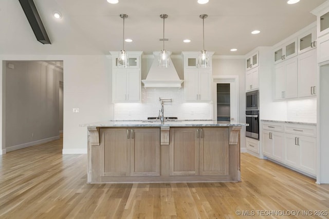kitchen featuring decorative backsplash, a kitchen island with sink, custom exhaust hood, light stone countertops, and light wood-type flooring