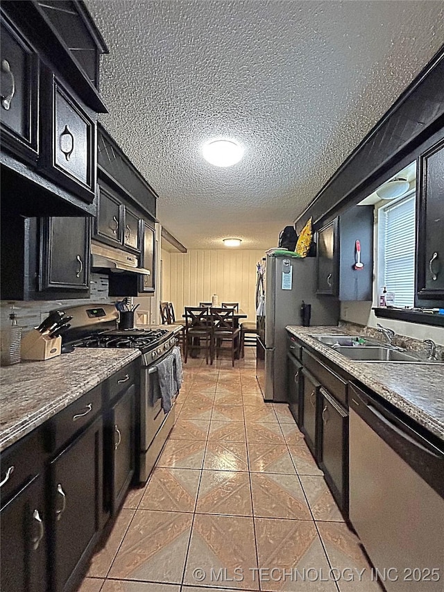 kitchen with a textured ceiling, appliances with stainless steel finishes, dark cabinets, under cabinet range hood, and light tile patterned flooring