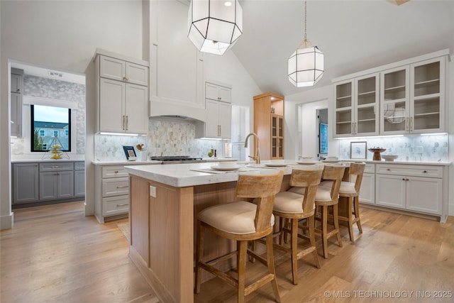 kitchen with glass insert cabinets, tasteful backsplash, a center island with sink, and light wood-type flooring