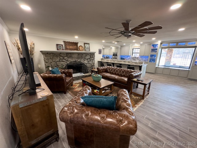 living area featuring a stone fireplace, light wood-style flooring, a ceiling fan, recessed lighting, and ornamental molding