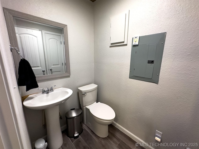 bathroom featuring wood finished floors, toilet, baseboards, electric panel, and a sink