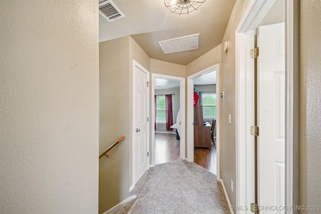 hall with visible vents, carpet flooring, a textured wall, and an upstairs landing