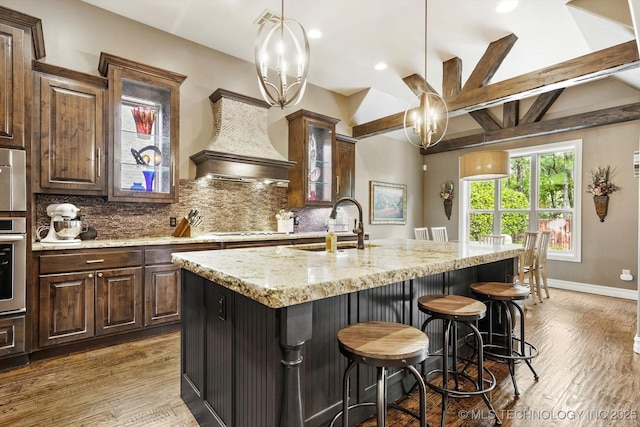 kitchen featuring light stone counters, wood finished floors, tasteful backsplash, a center island with sink, and glass insert cabinets