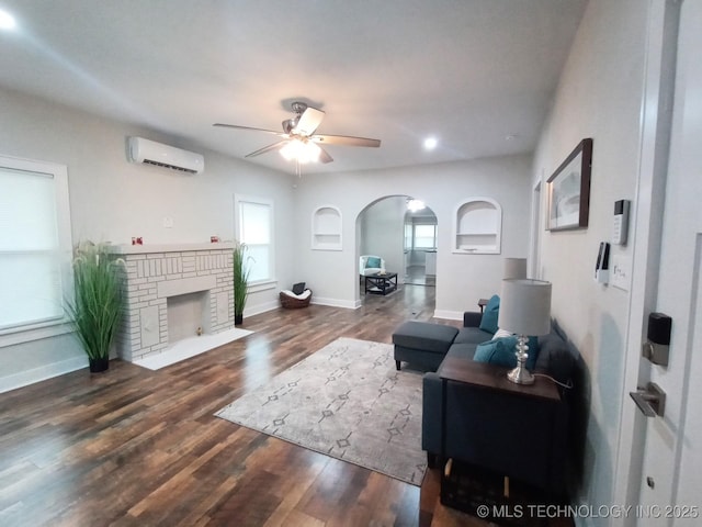 living room with a brick fireplace, an AC wall unit, arched walkways, a ceiling fan, and wood finished floors
