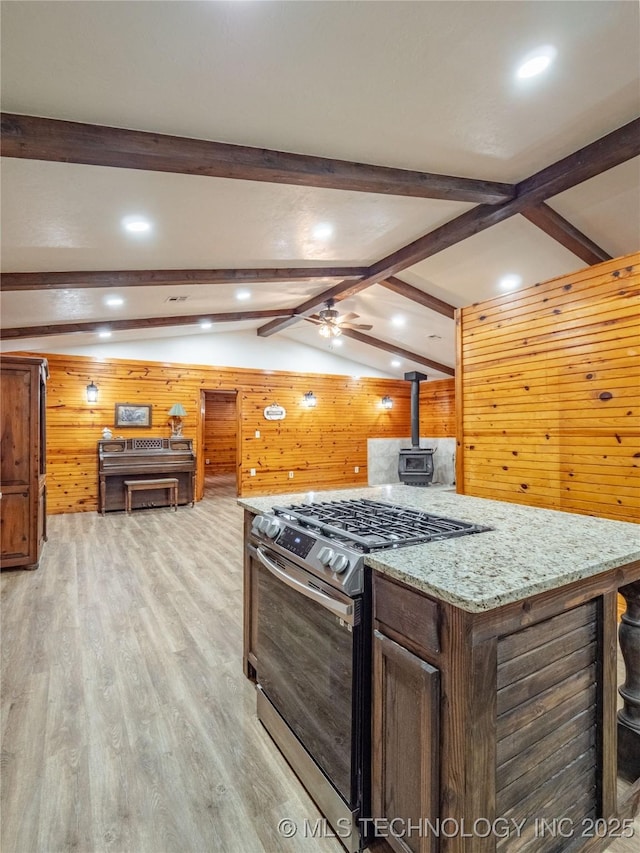 kitchen with wood walls, a wood stove, stainless steel gas stove, vaulted ceiling with beams, and light wood finished floors