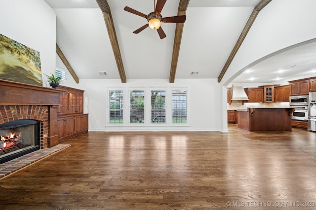 unfurnished living room with ceiling fan, a fireplace, beamed ceiling, arched walkways, and dark wood-style flooring