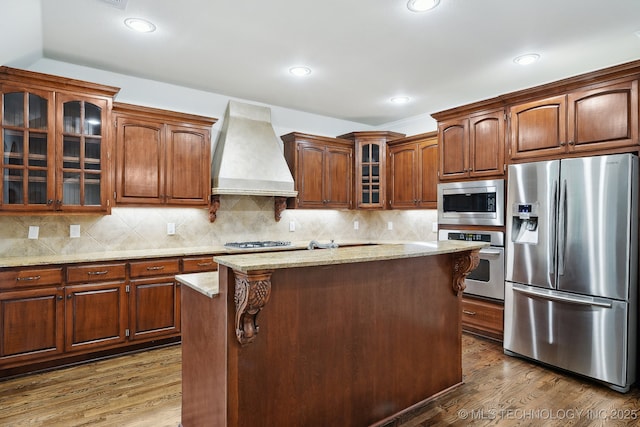 kitchen with appliances with stainless steel finishes, custom range hood, dark wood-type flooring, a center island, and light stone countertops
