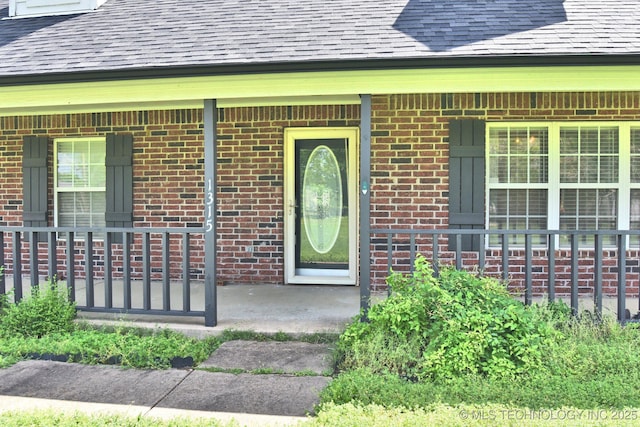 doorway to property with a shingled roof, a porch, and brick siding