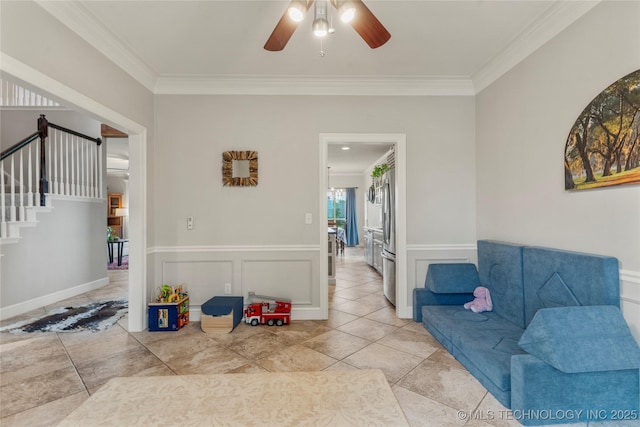 living room featuring ceiling fan, ornamental molding, stairway, and a wainscoted wall