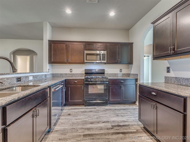 kitchen with a sink, stainless steel appliances, dark brown cabinetry, arched walkways, and light stone countertops