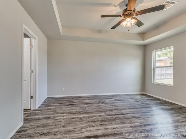empty room with a ceiling fan, a tray ceiling, baseboards, dark wood finished floors, and visible vents