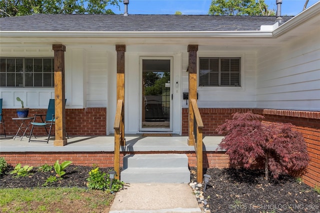 view of exterior entry featuring roof with shingles, covered porch, and brick siding