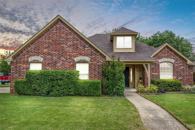 view of front facade with brick siding, a lawn, and roof with shingles