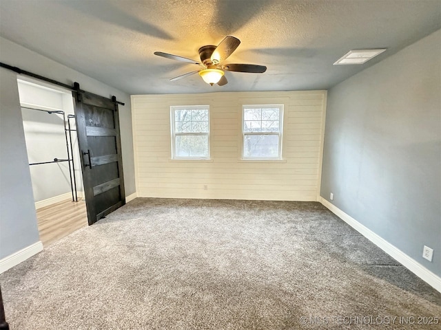 carpeted empty room featuring wooden walls, a barn door, a textured ceiling, a ceiling fan, and baseboards