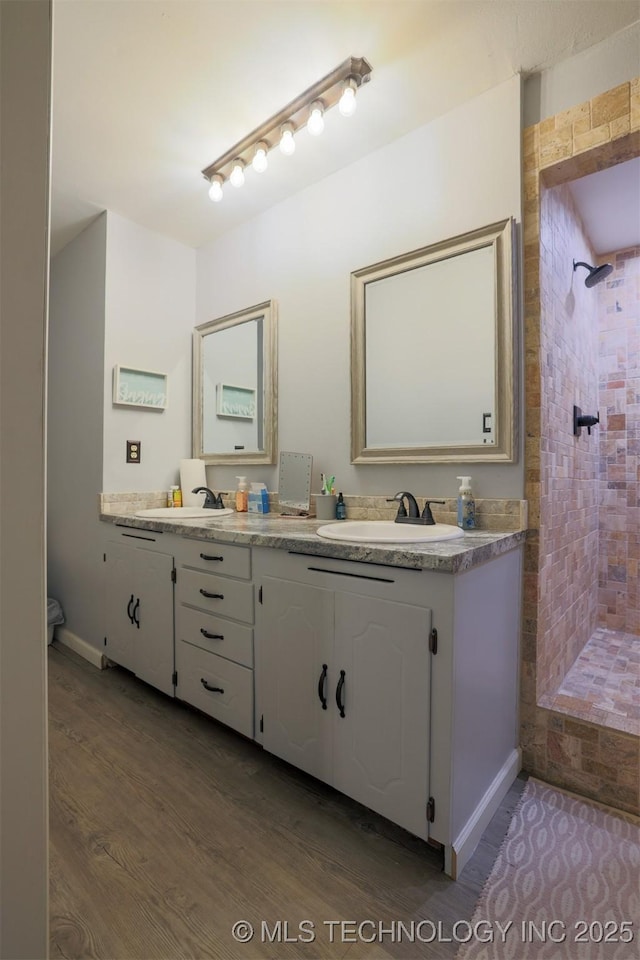 full bath with a sink, a tile shower, double vanity, and wood finished floors