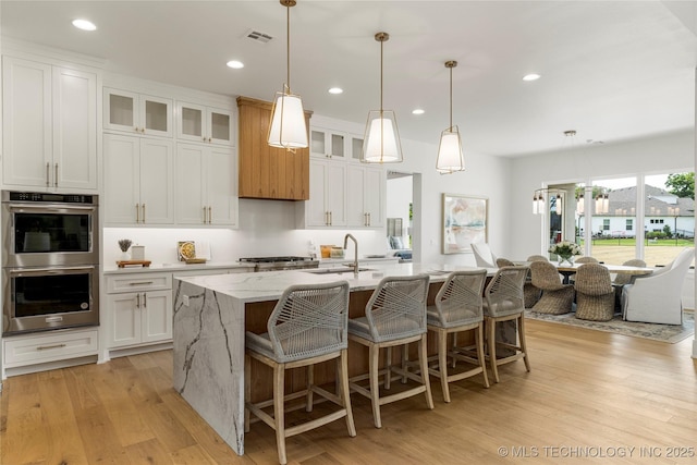 kitchen with a kitchen island with sink, stainless steel double oven, a sink, white cabinets, and light wood finished floors