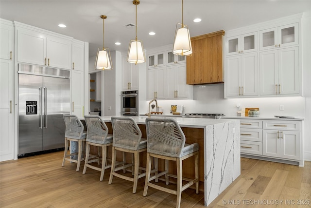 kitchen with a kitchen island with sink, white cabinetry, light wood-style flooring, appliances with stainless steel finishes, and decorative light fixtures