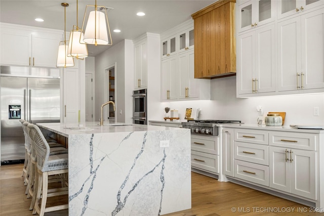 kitchen with an island with sink, white cabinets, light wood-style flooring, hanging light fixtures, and stainless steel appliances
