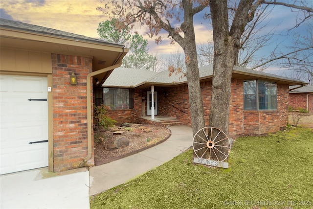 exterior entry at dusk with roof with shingles, an attached garage, a lawn, and brick siding