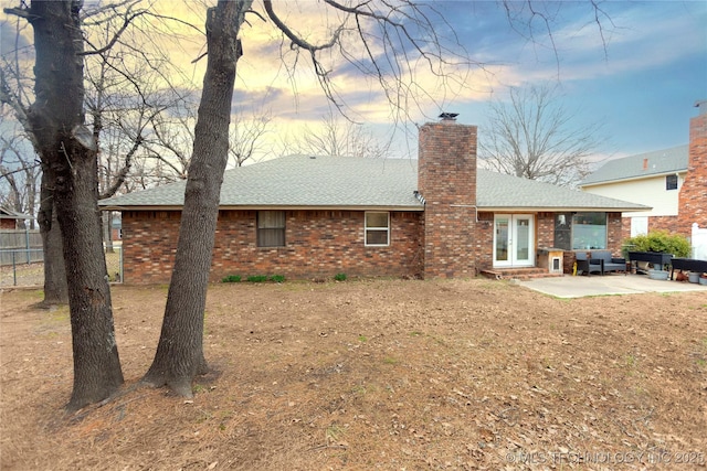 back of house at dusk featuring brick siding, a shingled roof, a chimney, and a patio