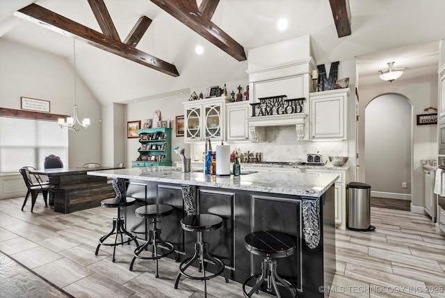 kitchen with light stone countertops, lofted ceiling with beams, a kitchen breakfast bar, arched walkways, and backsplash