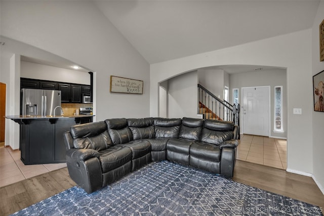 living room featuring stairs, arched walkways, high vaulted ceiling, and light wood-style floors
