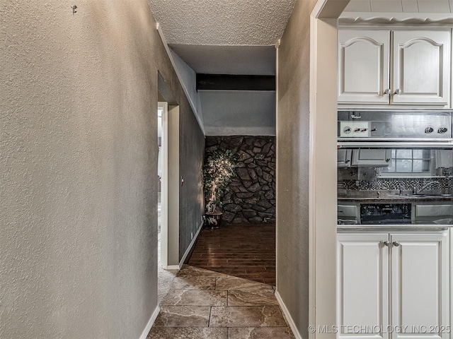 hall featuring baseboards, a textured ceiling, a textured wall, and stone finish flooring