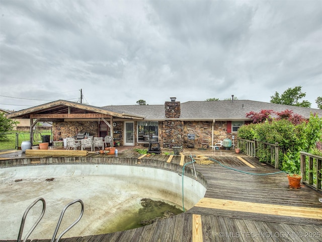 rear view of property featuring roof with shingles, a wooden deck, stone siding, and a chimney
