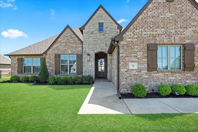 french country style house featuring brick siding, a shingled roof, stone siding, and a front lawn