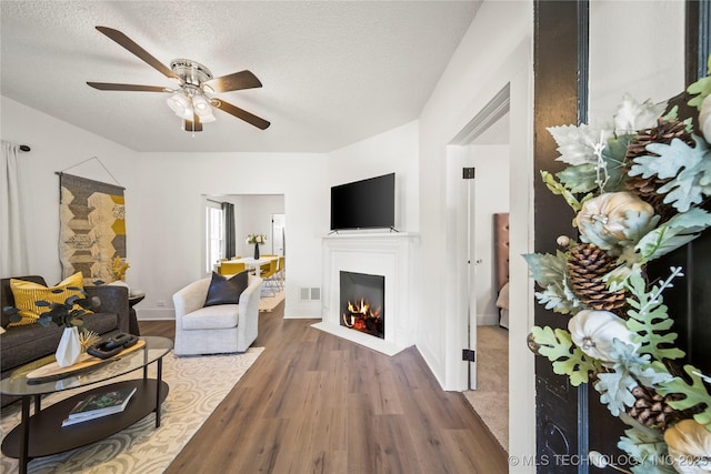 living area featuring a warm lit fireplace, a textured ceiling, a ceiling fan, wood finished floors, and visible vents