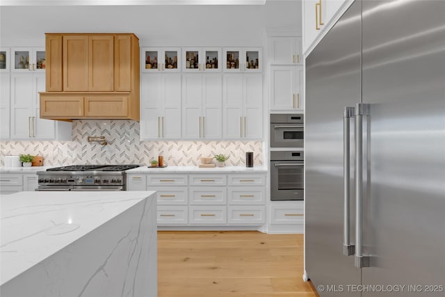 kitchen featuring decorative backsplash, glass insert cabinets, high quality appliances, white cabinetry, and light wood-style flooring