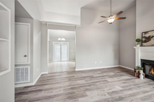 unfurnished living room featuring visible vents, wood finished floors, baseboards, ceiling fan with notable chandelier, and a warm lit fireplace