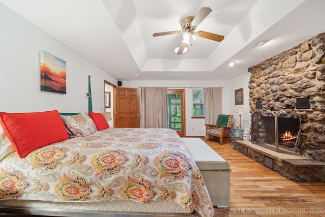 bedroom featuring access to outside, a raised ceiling, a stone fireplace, a ceiling fan, and wood finished floors