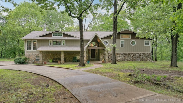 view of front of house with covered porch, a front yard, stone siding, a shingled roof, and driveway