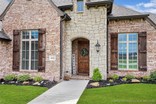 entrance to property featuring a yard, brick siding, a shingled roof, and stone siding