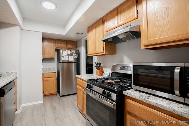 kitchen with appliances with stainless steel finishes, light stone counters, a raised ceiling, light wood-style floors, and under cabinet range hood